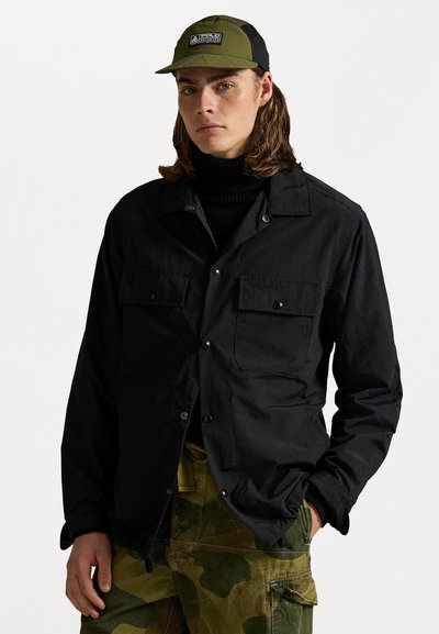 Sort letvægtsjakke med to brystlommer og knaplukning, båret over en sort turtleneck, parret med camo cargo-bukser.