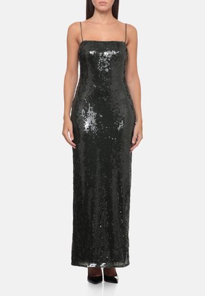 Donna che indossa un abito lungo nero con paillettes, con spalline sottili e scarpe nere con tacco alto a punta, in piedi davanti a uno sfondo semplice.