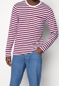 Mann in einem langärmligen, maroon- und weißgestreiften Hemd mit Brusttasche, kombiniert mit blauen Jeans, Hand in der Tasche.