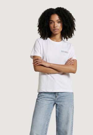 Mujer con cabello rizado, vistiendo una camiseta blanca con un pequeño texto en el pecho y jeans azul claro, de pie con los brazos cruzados.