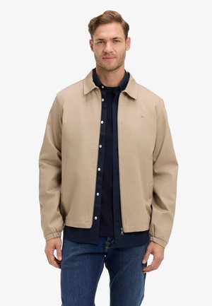 Jeune homme portant une veste beige par-dessus une chemise bleu marine à boutons et un jean foncé, debout devant un fond blanc uni.