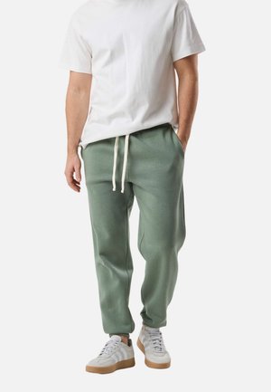 Mężczyzna ubrany w prostą białą koszulkę, szare zielone spodnie dresowe ze ściągaczem i białe trampki z gumową podeszwą, stojący na tle białego tła.