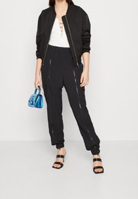 Svart bomberjacka över en vit topp med snörning framtill, ihop med svarta tapered byxor med dragkedjor och svarta sandaler med remmar. En liten blå handväska fulländar looken.