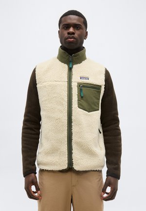 Jaunas vyras dėvi smėlio spalvos vilnonį liemenę su žalia krūtinės kišene ir rudomis ilgomis rankovėmis, stovintis priešais paprastą šviesią foną.