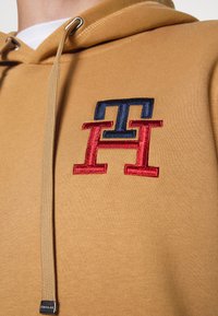 Senapsgul hoodie med en mjuk textur, med en mångfärgad broderad logotyp i marinblått och rött på bröstet, och en huva med dragsko.