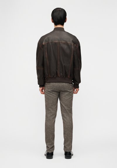 Άνδρας που στέκεται γυρίζοντας την πλάτη, φορώντας ένα σκούρο καφέ δερμάτινο μπουφάν, γκρι υφασμάτινο παντελόνι και μαύρα επίσημα παπούτσια, πάνω σε λευκό φόντο.