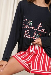 Fekete, hosszú ujjú póló "Santa Baby" felirattal és nyalóka díszítéssel; piros-fehér csíkos rövidnadrág fehér masnival a derekán.