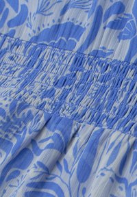 Tissu froncé bleu et blanc avec motifs floraux et feuillage, montrant une couture détaillée et une texture plissée.