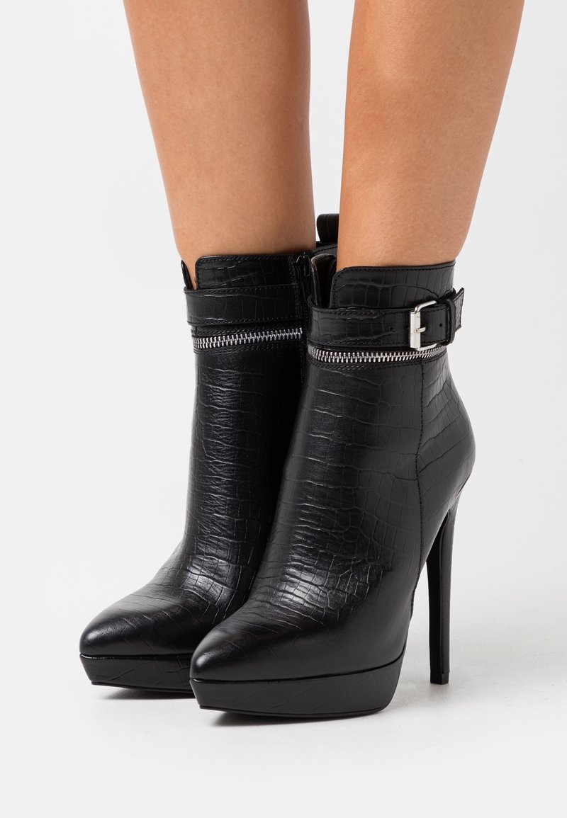 Bottes à talons hauts noires jusqu'à la cheville avec bouts pointus, texture croco, détail de fermeture éclair argentée et brides réglables avec boucles portées sur des jambes nues.