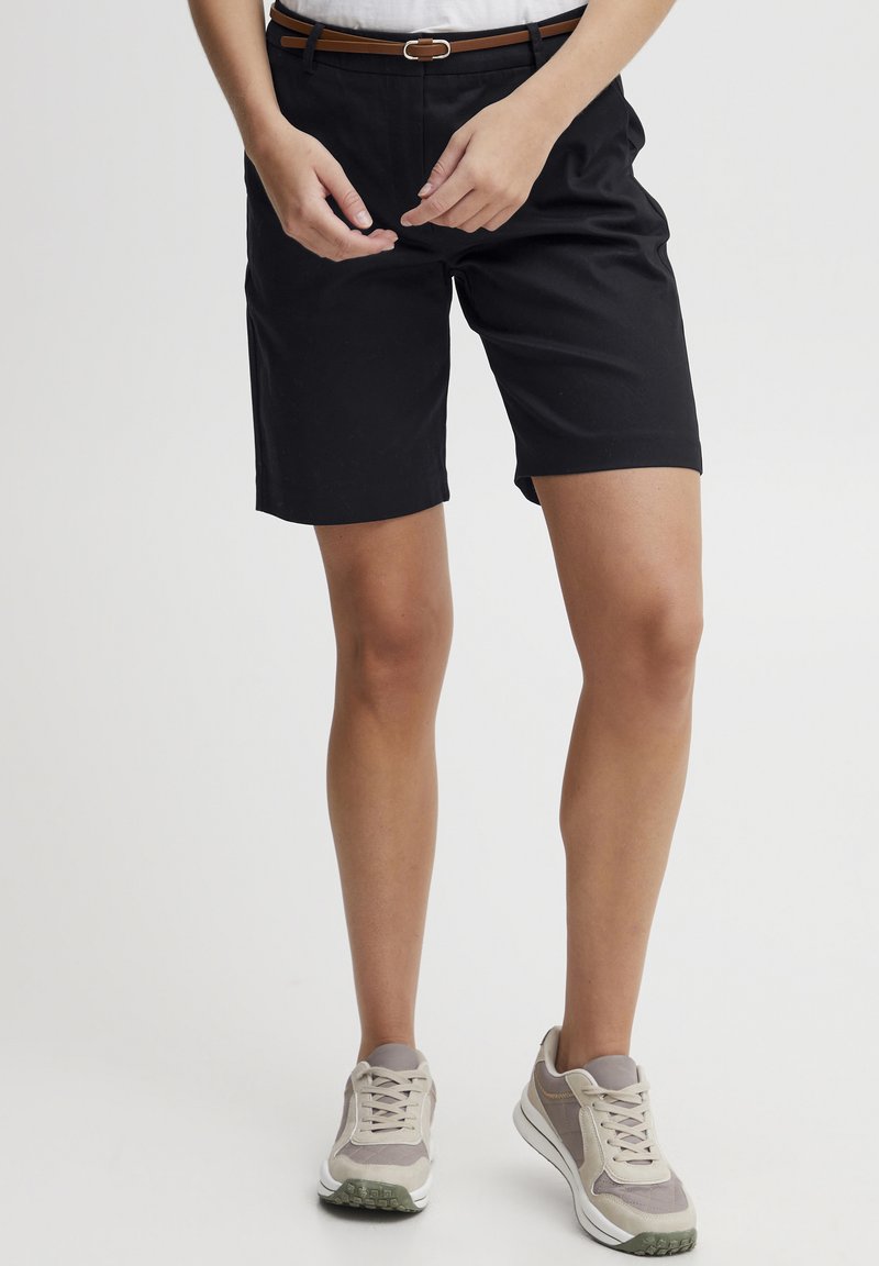 Shorts noirs au design ajusté, avec une texture lisse et une ceinture marron. Associés à des baskets grises clair et une chemise blanche.