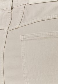 Beige Denimstoff von Hosen, mit einer Vordertasche, sichtbaren Nähten und einer glatten, strukturierten Oberfläche. Detail der Gesäßtasche nicht gezeigt.