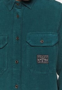 Tealfärgad kordflorsskjorta med vertikala ribbor, svarta knappar, två bröstfickor och en broderad logotyp med "KAOTIKO."