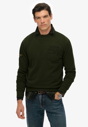 Mand iført en mørkegrøn sweatshirt over en grønternet skjorte med blå jeans, ur på håndleddet, står med den ene hånd i lommen.