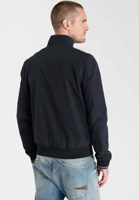 Veste bleu marine foncé avec un col haut, un ourlet élastiqué et des poignets. Présente des détails de couture subtils et une coupe décontractée. Portée avec un jean bleu clair.