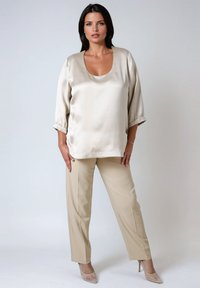 Woman with dark hair wearing beige satin blouse, matching beige straight pants, and nude high heels standing against plain gray background.