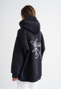 Übergroßer schwarzer Hoodie mit einem weißen abstrakten Gesichtsmuster auf der Rückseite, ausgestattet mit einer Kordelzugkapuze sowie gerippten Bündchen und Saum.