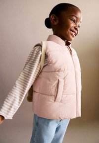 Svetloružová prešívaná vesta s vysokým golierom, predným zipsom a bočnými vreckami, nosená cez pruhovanú blúzku s dlhým rukávom. Mäkká textúra.