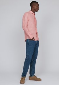 Svetlo roza dolg rokav, s gumbi zapet s teksturirano tkanino, v kombinaciji z modrimi jeans hlačami in bež čevlji. Model stoji v profilu.