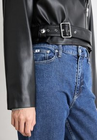 Giacca in pelle nera con cintura larga e fibbia d'argento, abbinata a jeans in denim blu con effetto sbiadito delicato e dettaglio del logo sulla vita.