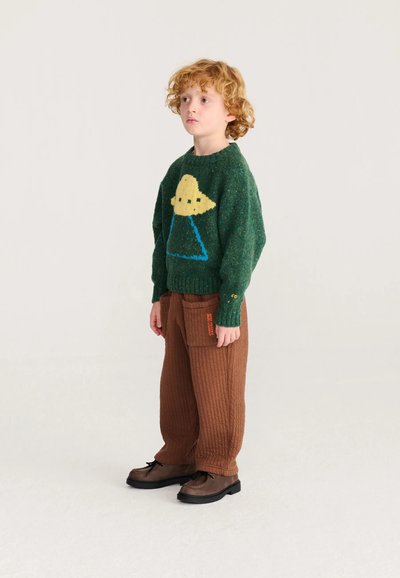Enfant aux cheveux roux bouclés portant un pull vert avec un motif OVNI jaune, un pantalon marron texturé et des chaussures marron foncé, debout devant un fond uni.
