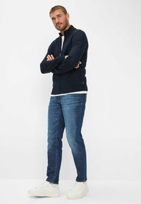 Marine blauwe ritstrui met geribde manchetten, gedragen over een wit shirt, gecombineerd met blauwe jeans en witte sneakers. Model staat met gekruiste armen.