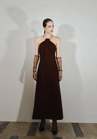 Brązowa sukienka na halter z płynącą sylwetką. Posiada przezroczyste, siateczkowe długie rękawy oraz długość do kostek. Gładki materiał z minimalnymi zdobieniami.