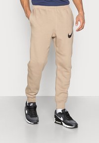 Beige tepláky s elastickým pasem a nohavicemi, s černým logem Nike na levém stehně, obuté s černobílými teniskami Nike.