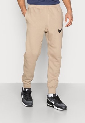 Pantalons de jogging beiges avec taille élastique et poignets, arborant un logo Nike noir sur la cuisse gauche, portés avec des baskets Nike noires et blanches.