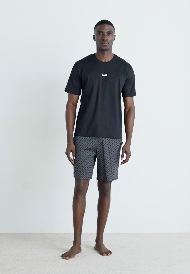 T-shirt noir à manches courtes avec un petit logo, associé à un short gris à motifs. Tissu léger, coupe décontractée. Le modèle est pieds nus.