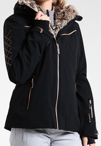 Schwarze Jacke mit gefütterter Kapuze aus Fell, roségoldenen Akzenten, Reißverschlusstaschen und diagonalen Nähten an den Ärmeln. Der Stoff wirkt glatt und isoliert.