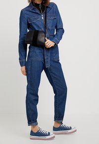 Tuta in denim con zip frontale, maniche lunghe e due tasche sul petto. Abbinata a sneakers in tela blu e una piccola borsa nera.