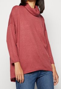 Cowl-Neck, langärmliges Oberteil in einer strukturierten Rostfarbe. Lockere Passform mit leicht gewelltem Saum und gerippten Bündchen. Getragen mit blauen Jeans.