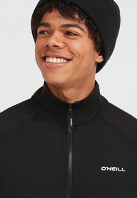 Zwarte ritsjack met hoge kraag, gemaakt van rekbaar materiaal, met een witte "O'NEILL" logo op de linkerkant van de borst en een zwart geribbeld beanie.