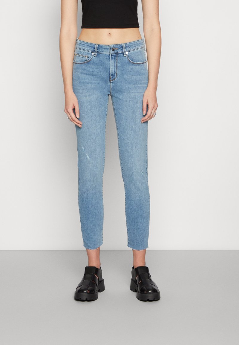 Jeans de ganga elásticos em azul claro com um corte slim, apresentando uma cintura de altura média, um desvanecimento subtil e um comprimento até ao tornozelo. Sapatos pretos chunky completam o visual.