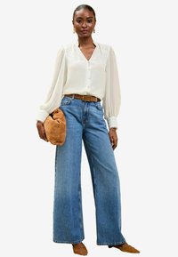 Blusa bianca a maniche lunghe con bottoni e dettagli in pizzo, abbinata a jeans blu a gamba larga. Cintura beige e borsa beige rotonda come accessori.