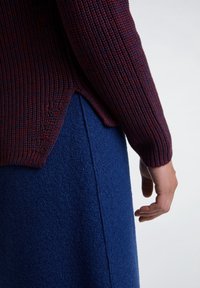 Maglione a maglia di un rosso scuro con una texture a coste abbinato a una gonna di lana blu. Primo piano della manica e della mano.