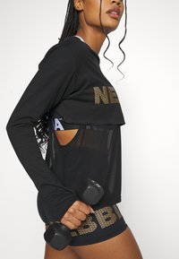 Svart sportslig langermet topp med mesh-overlegg og gulltrykt tekst, sammen med tettsittende svarte shorts med dristig skrift. Holder en hantel.