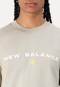 Gaiši pelēks īsais krekls ar īsām piedurknēm, uz kura ir uzraksts "NEW BALANCE" baltā krāsā un dzeltenais logo. Mīksta tekstūra, apaļa kakla izgriezums un brīvs piegriezums.