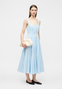 Frau trägt ein ärmelloses Midi-Kleid mit hellblauen und weißen Streifen, hält eine beige Clutch und trägt schwarze Ballerinas sowie goldene Creolen.