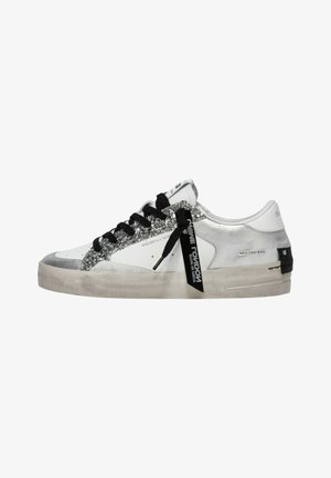 Sneaker bianca basse con dettagli glitter argento, lacci neri, etichetta nera con marchio e suola beige leggermente consumata.