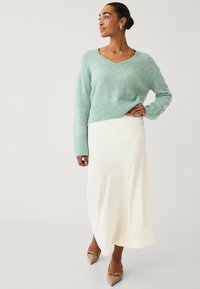 Suéter de punto verde menta con escote en V, combinado con una falda maxi de satén blanca y unos tacones beige de punta con un acento de herrajes decorativos.