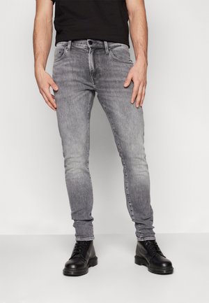 Mann trägt ausgewaschene graue Skinny-Jeans und schwarze Schnürstiefel, steht vor einem einfachen hellen Hintergrund.