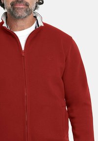 Chaqueta roja con cremallera y cuello alto, con un forro interior blanco. Fabricada en tela suave, cuenta con dos bolsillos laterales y un discreto detalle de logo.