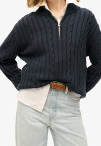 Marineblauer Pullover mit Zopfmuster und Reißverschluss am Kragen, strukturierten Streifen und geripptem Kragen, kombiniert mit einem weißen Hemd und hellblauen Jeans. Getragen mit einem geflochtenen braunen Gürtel.