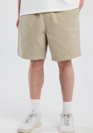 Pantaloni corti in cotone beige con taglio dritto e tasche laterali, abbinati a scarpe da ginnastica bianche e calzini a coste. Texture liscia, design casual.