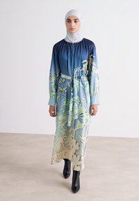 Ein langes, fließendes Kleid mit einem Farbverlauf in Blau und floralen Mustern, gerafftem Ausschnitt und einem passenden Gürtel. Kombiniert mit schwarzen Stiefeletten.