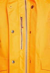 Giacca impermeabile gialla con una texture liscia, zip frontale con denti in metallo, bottoni a pressione e tasche laterali.