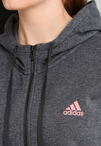 Sweat à capuche zippé gris foncé en tissu doux. Dispose d'une fermeture éclair noire et d'un logo Adidas rose sur la poitrine. Comprend une capuche et des cordons de serrage.