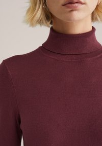 Bordo turtleneck megztinis su rausvu tekstūru, prigludusia siluetu ir aukštu kaklu, demonstruojantis sklandų audinio apdailą.
