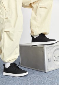 Personne portant un pantalon beige et des baskets noires, avec un pied sur une boîte grise métallisée sur un sol en moquette bleue.